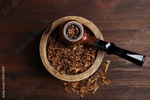 Smoking pipe and bowl of dry tobacco on wooden table, top view