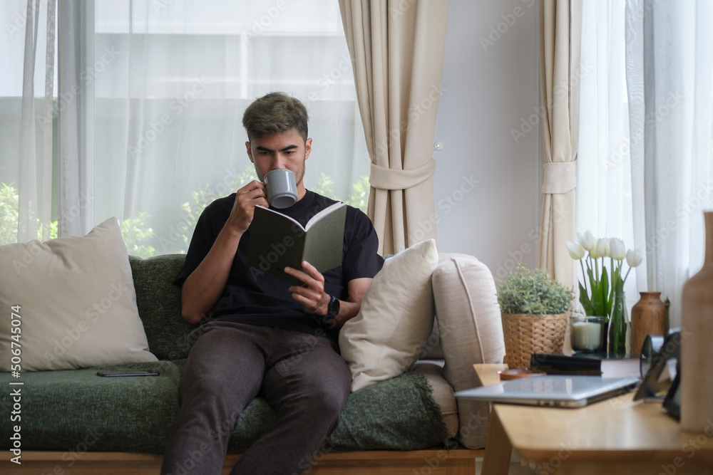Positive young man reading book and drinking coffee while sitting on sofa at home. Positive young man reading book and drinking coffee while sitting on sofa at home.