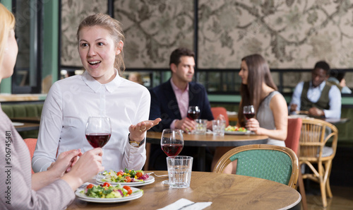 Happy attractive girl emotionally discussing with female friend while dining in cozy restaurant