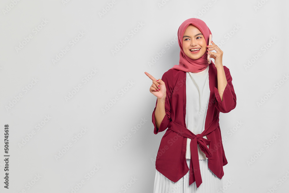 Cheerful beautiful Asian woman in casual shirt talking on mobile phone and pointing fingers at copy space isolated over white background