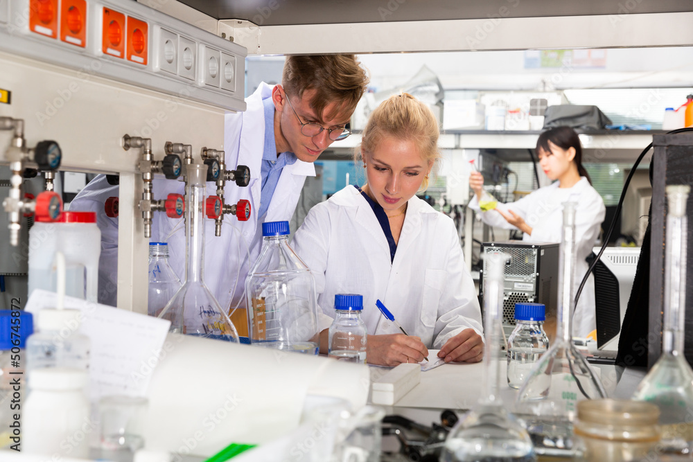 Fototapeta premium Two students of Faculty of Chemistry performing experiments in university laboratory, recording results in workbook
