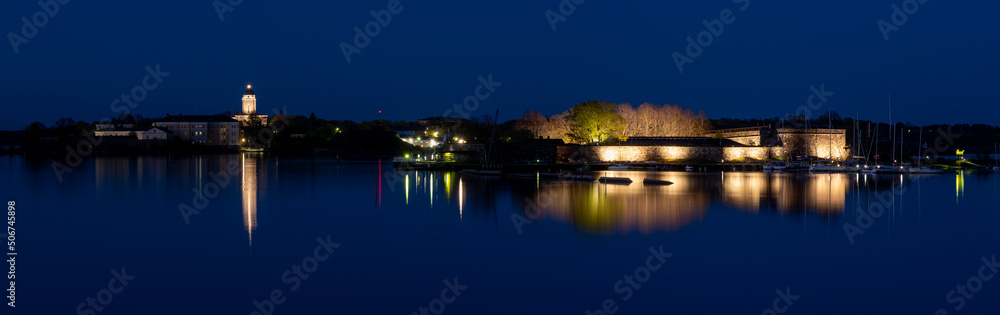 Fototapeta premium Panoramic view of Suomenlinna island. Illuminated buildings cast reflections on the calm water.
