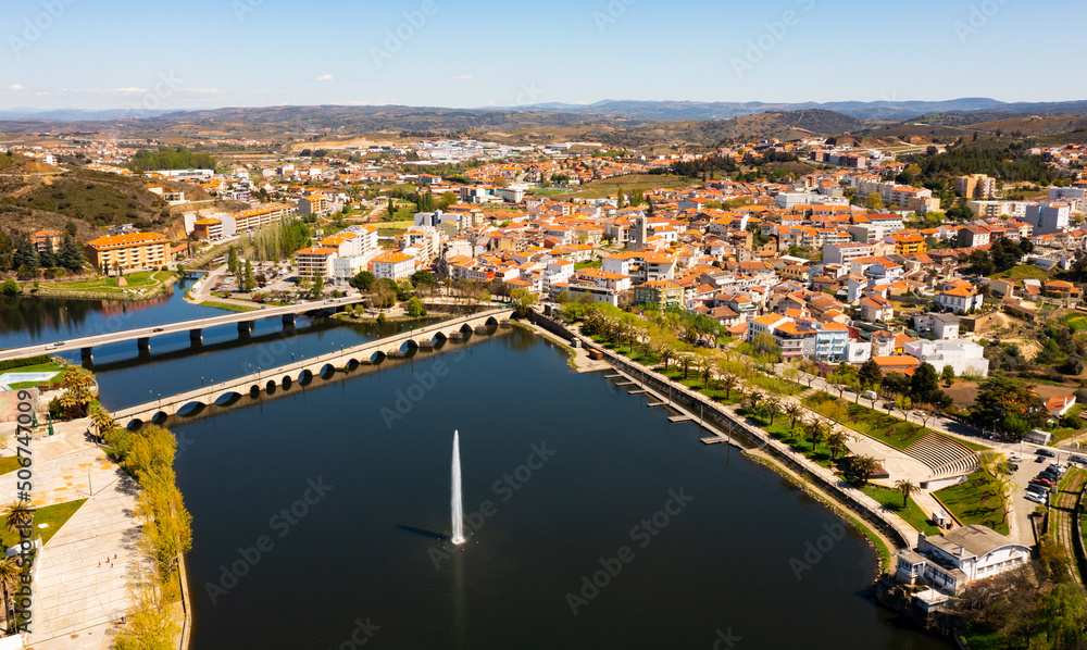 Fototapeta premium Picturesque aerial view of Mirandela city with residential houses with brownish tiled roofs, bridges crossing Tua river and water fountain on spring day, Braganca, Portugal