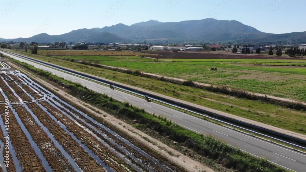 Aerial View Of Agricultural Fields, Mountains And Two Cyclists Riding On The Highway Aerial View Of Agricultural Fields, Mountains And Two Cyclists Riding On The Highway