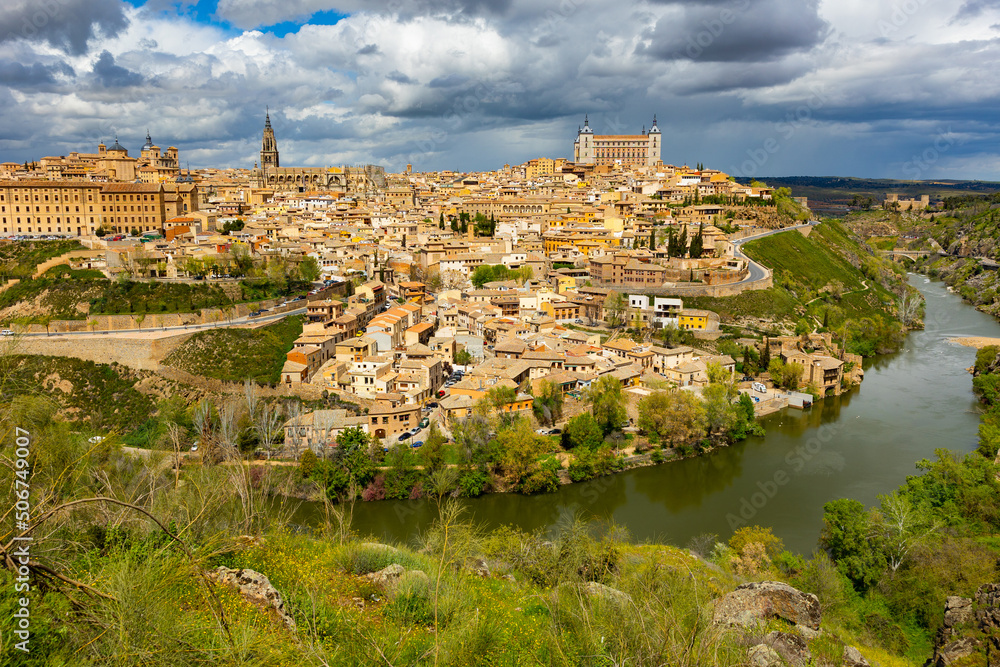 Picturesque view of Toledo cityscape on green hilly banks of Tagus river in early spring overlooking Gothic bell tower of ancient Cathedral and fortified Alcazar castle, Spain