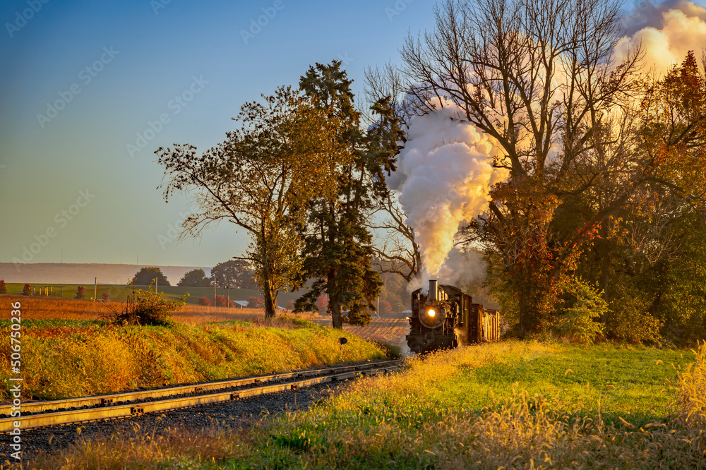 A View of an Antique Freight Steam Train Blowing Smoke Approaching Thru Trees in Late Afternoon