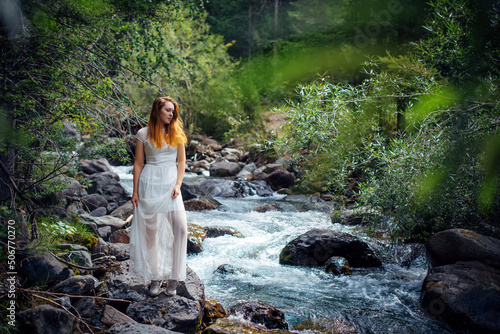Slender long-haired brunette in white dress posing against small mountain river and green trees. Beautiful young woman walking along the forest stream shore.