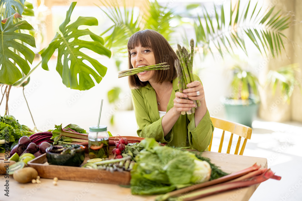 Obraz premium Portrait of a young cheerful woman holds a bunch of asparagus while sitting by the table full of fresh vegetables, fruits and greens. Healthy eating and lifestyle concept