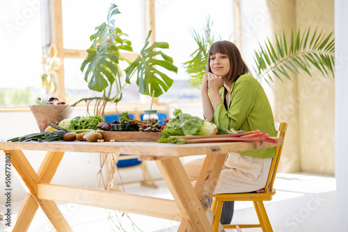 Portrait of a young cheerful woman holds a bunch of asparagus while sitting by the table full of fresh vegetables, fruits and greens. Healthy eating and lifestyle concept