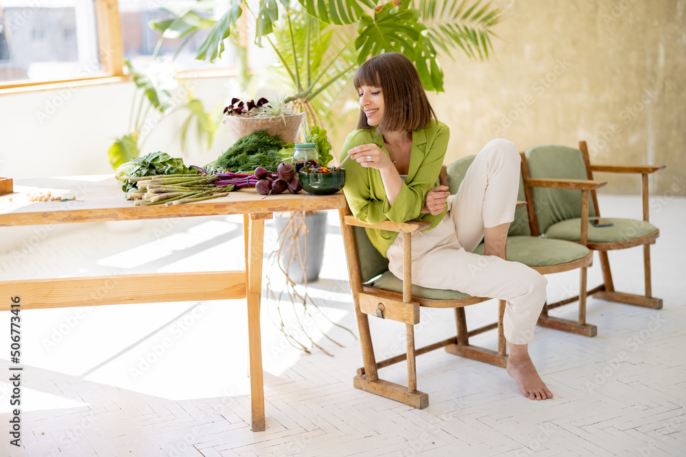 Young woman eats vegetarian lunch in bowl while sitting on chair near table with lots of fresh food ingredients in room with green plants. Healthy lifestyle concept