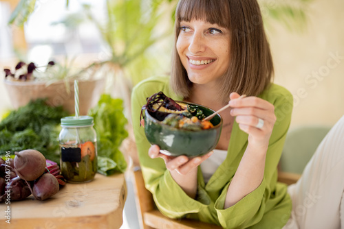 Young cheerful woman eats vegetarian lunch in bowl, sitting by the table full of fresh food ingredients indoors. Healthy lifestyle and wellness concept