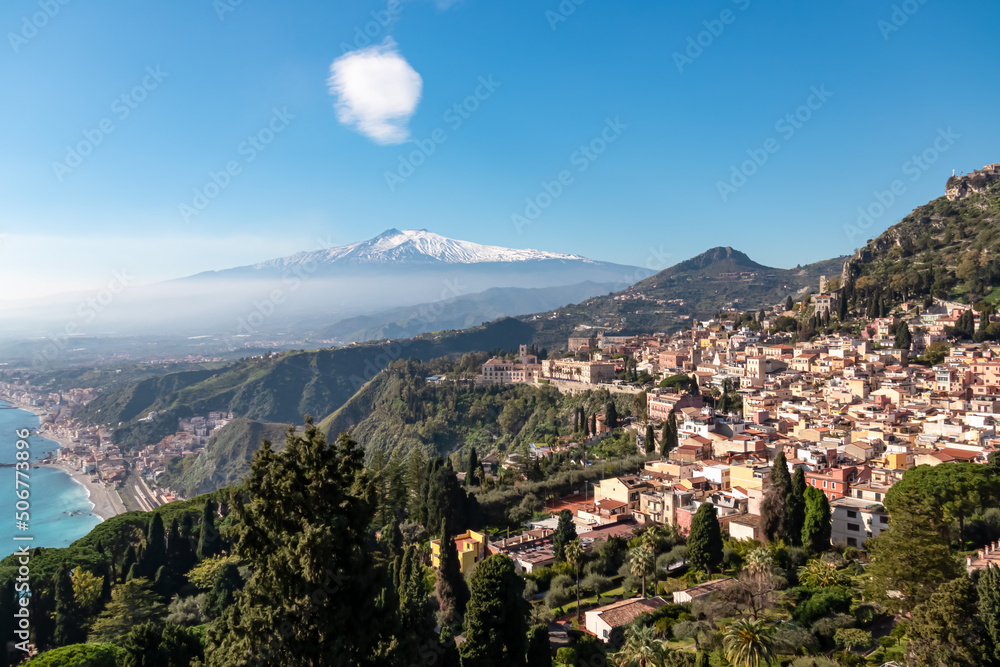 Panoramic view on snow capped Mount Etna volcano and Mediterranean coastline on sunny day seen from coastal hilltop town Taormina, island Sicily, Italy, Europe, EU. Vacation tourism travel destination