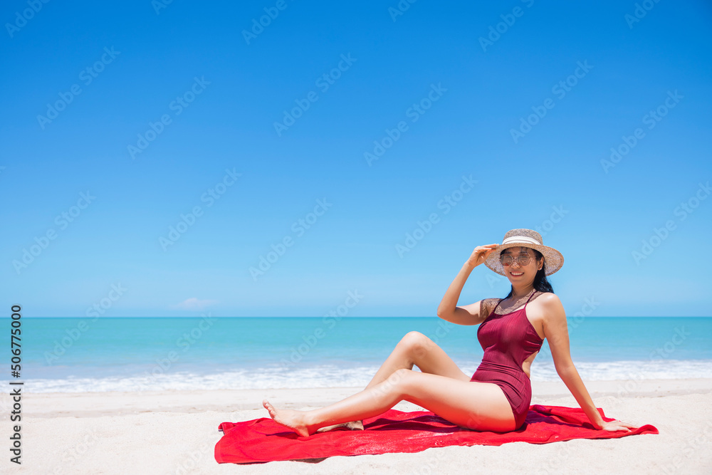 Young women in bikini and straw hat sit on tropical beach enjoying looking view of beach ocean on hot summer day. Blue sea in background. Khao Lak, Phang Nga, Thailand. Summer vacation concept