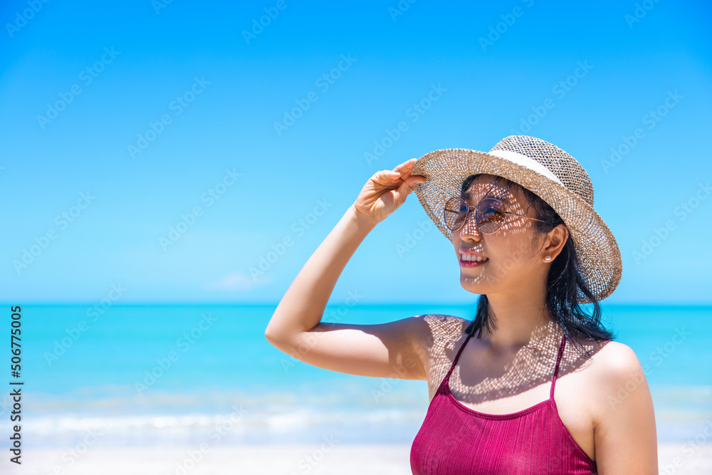 Young women in bikini and straw hat stand on tropical beach enjoying looking view of beach ocean on hot summer day. Blue sea in background. Khao Lak, Phang Nga, Thailand. Summer vacation concept