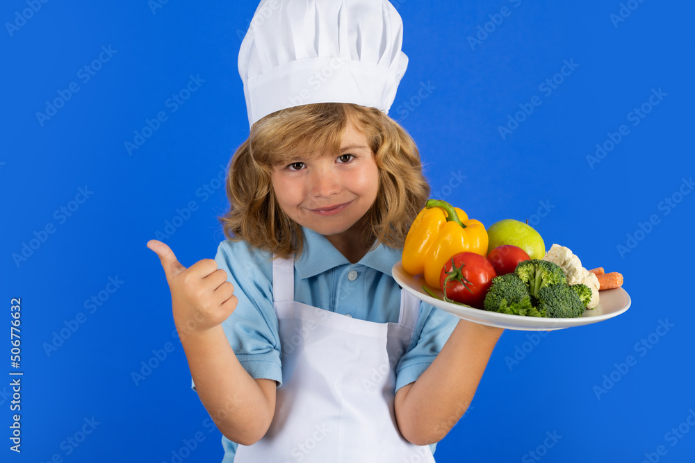 Child chef dressed cook baker apron and chef hat hold plate with vegetables isolated on studio background. Healthy nutrition kids food. Child chef dressed cook baker apron and chef hat hold plate with vegetables isolated on studio background. Healthy nutrition kids food.