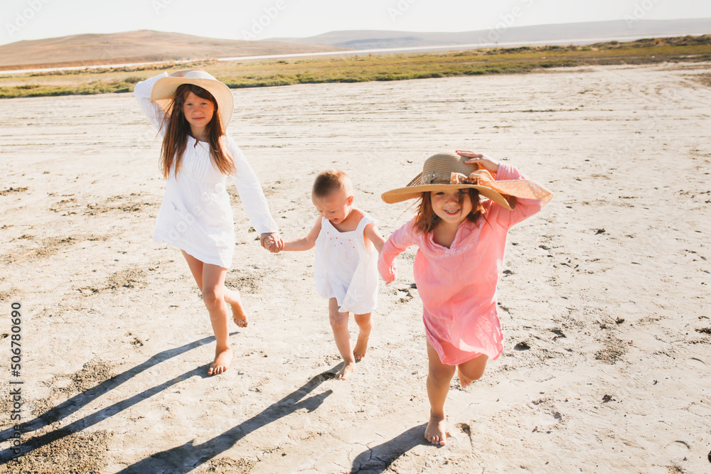 three children hold hands and run together deserted beach, dried-up mud lake. Summer children's recreation in nature. Children in tunic dresses from the sun, three children hold hands and run together deserted beach, dried-up mud lake. Summer children's recreation in nature. Children in tunic dresses from the sun,