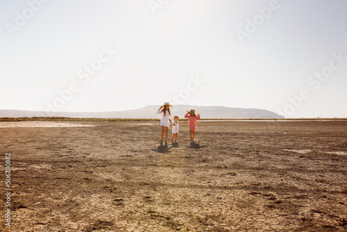 three children hold hands and run together deserted beach, dried-up mud lake. Summer children's recreation in nature. Children in tunic dresses from the sun,