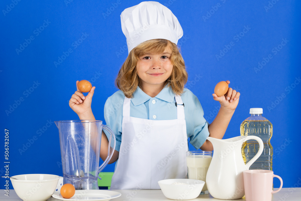 Portrait of a 7, 8 years old child in cook cap and apron hold eggs making fruit salad and cooking food in kitchen. Cute little blonde happy smiling chef. Portrait of a 7, 8 years old child in cook cap and apron hold eggs making fruit salad and cooking food in kitchen. Cute little blonde happy smiling chef.