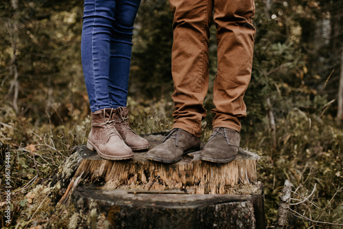 A couple is standing on a wooden trunk and their boots and jeans are on the photo. Close up shot of their trousers and brown footwear. Outdoor lifestyle in the forest. 