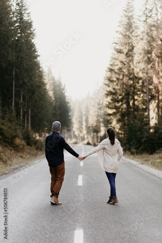 A couple in love is walking on an epmty road in the forest. Holding hand and keeping distance as they walk. Good relationship, active lifestyle. Together on the way. They face the distance. 