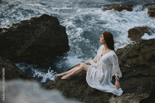 A woman in a white wedding dress sits on a rocky stone by the ocean just