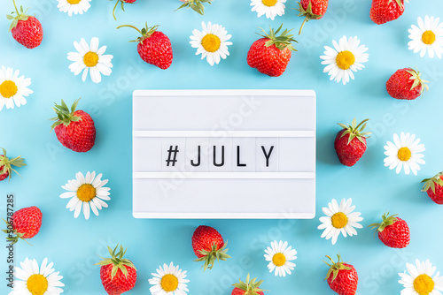 Summer month July text on light box and strawberry, chamomile flowers on blue background. Creative concept Hello July. Top view, Flat lay, greeting card