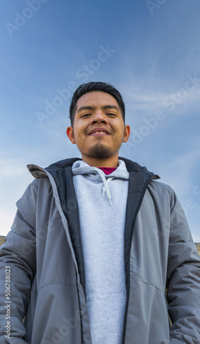 waist up image of a smiling young man with originarie mexican people features standing on a street with a clear sky behind him
