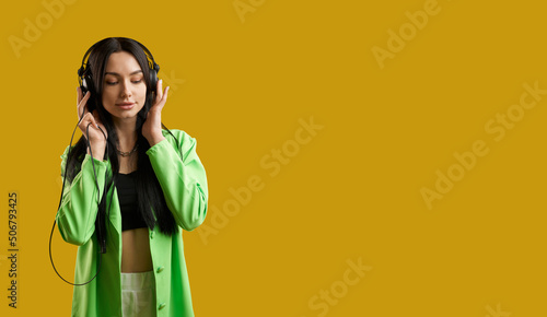 Front view of girl wearng green suit, standing, listening to music. Pretty brunette woman with long hair enjoying music with closed eyes, smiling. Concept of modern life. photo