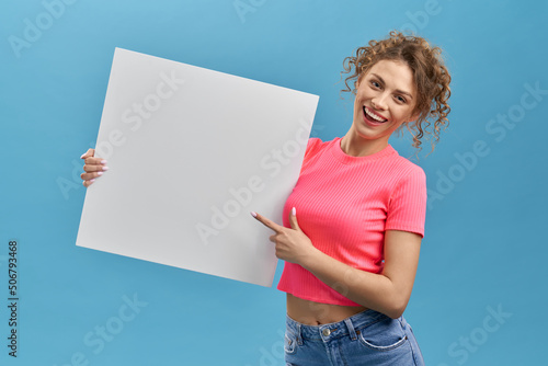 Front view of curly slim girl smiling, holding white board. Young blonde female standing, pointing by finger, wearing pink T-shirt and jeans. Isolated on blue studio background.