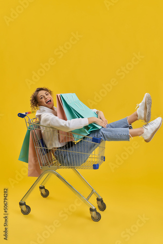 Side view of girl with curly hair doing shopping. Young female sitting in shopping cart, holding, hugging packages, looking at camera. Isolated on yellow studio background.