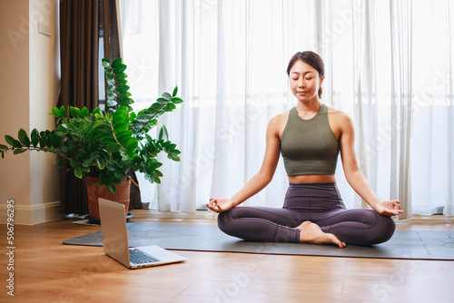 Asian woman practicing yoga from yoga online course on a video conference on a laptop at home.