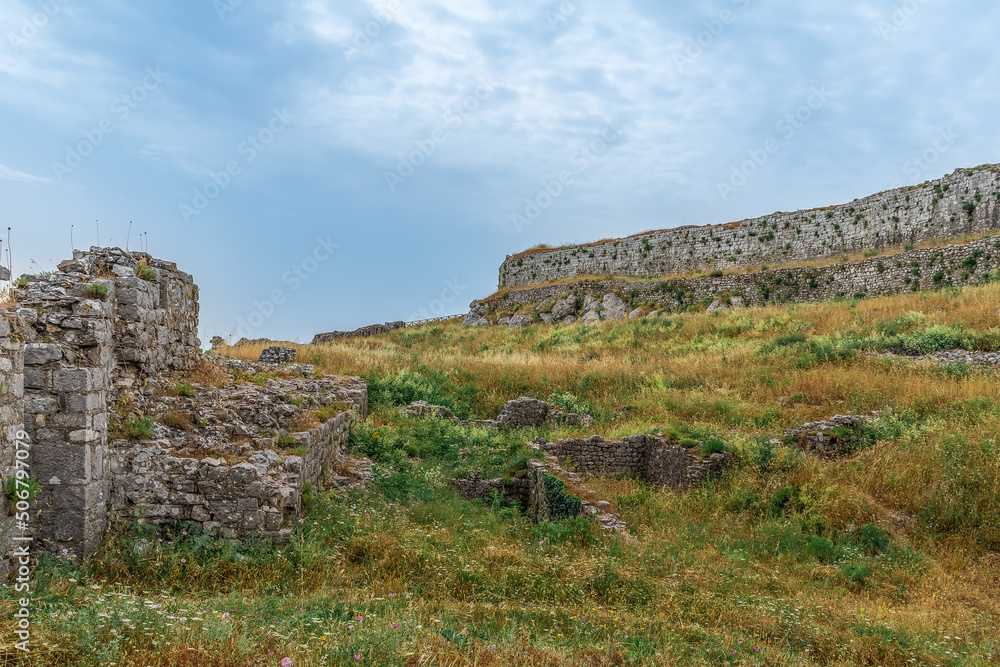 Meadow with yellow-green dry grass among the ruins of Rozafa Castle, Albania. Remains of stone fortification walls among the lawn against the blue sky