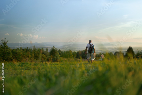 Back panoramic view of man with gray hair going forward alone in woods. Male with rucksack walking, hiking, traveling, enjoying scenery in hills, Concept of tourism.