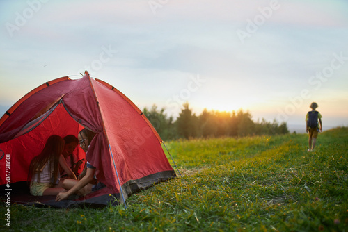 Panoramic view of red tent with friends inside, one tourist going alone forward by road. Group of people at sunset hiking, talking. Concept of mount traveling, active recreation. 