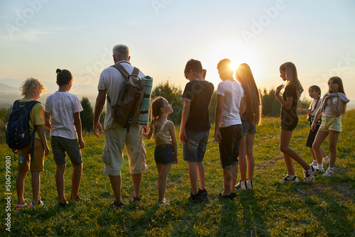 Back view of old man with kids standing in row in hills. Children with male with gray hair wearing rucksack traveling, hiking together, having break at sunset. Concept of active rest.