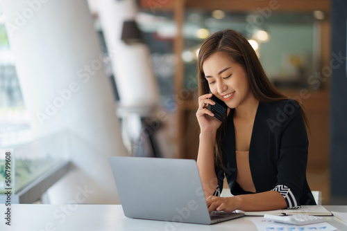 Happy secretary Asian business woman charming sitting smiling and talking on the smartphone in the office.