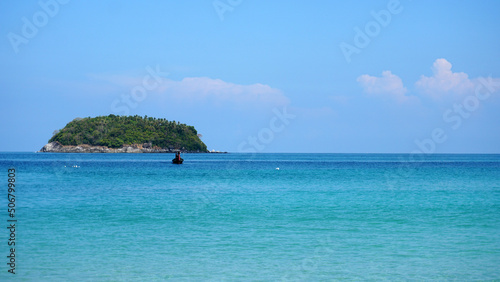 A lonely island in the sea. A Thai fishing boat floats nearby. Clear blue water, snow-white sand. Small clouds in the sea. Palm trees and trees grow on the island. Beautiful Kata beach, Phuket