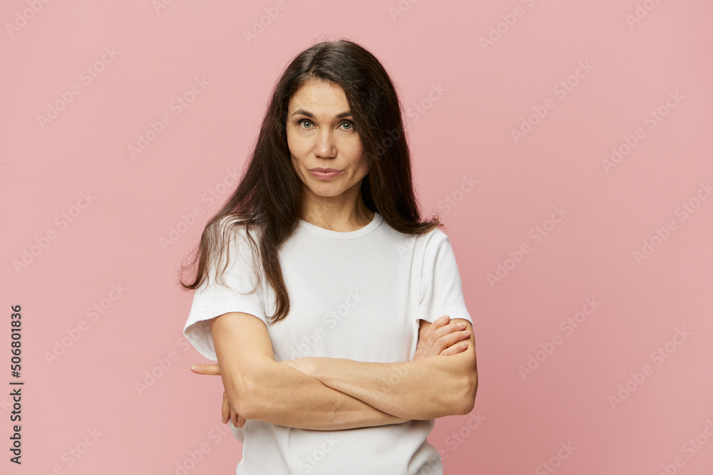 Obraz premium portrait of a beautiful, charismatic, attractive woman with dark hair color with arms crossed on her chest in a white cotton tank top on a pink background, with empty space