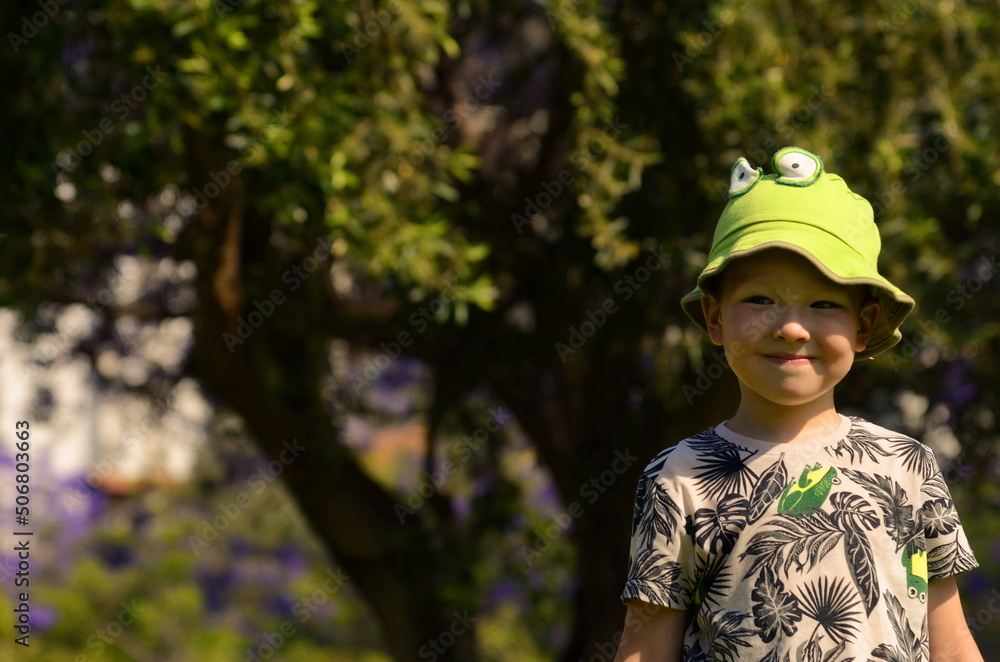 Positive smiling boy 5 years old, portrait against the background of blooming jacaranda, purple flowers. A child in a panama hat with a frog. Concept: good mood, children's joy, day off, botan
