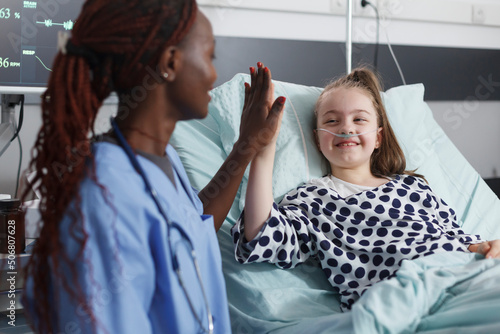 Pediatric hospital nurse highfive sick child while in patient treatment ward room. Blessed cheerful young patient highfive medical staff while in medical examination room. photo