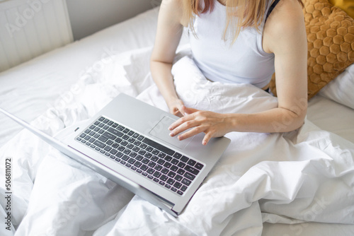 Happy casual beautiful woman working on a laptop sitting on the bed at the morning in the house. Working at home  freelance concept.