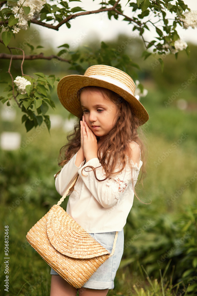 Beautiful sad girl in a straw hat against the backdrop of flowering gardens