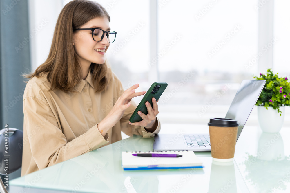 Young woman working from home using phone and computer, woman's hands using smart phone in interior, at home workplace using technology Young woman working from home using phone and computer, woman's hands using smart phone in interior, at home workplace using technology