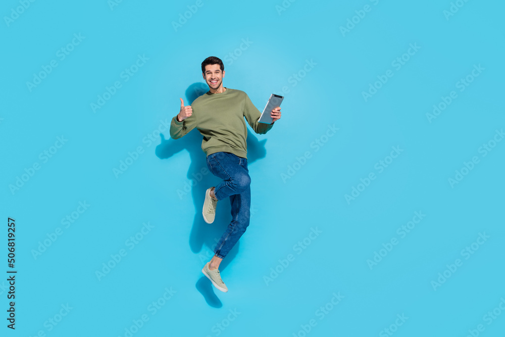 Full body photo of hooray young brunet guy jump hold tablet thumb up wear shirt jeans sneakers isolated on blue background Full body photo of hooray young brunet guy jump hold tablet thumb up wear shirt jeans sneakers isolated on blue background