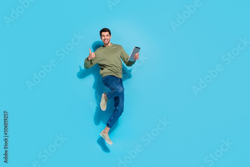 Full body photo of hooray young brunet guy jump hold tablet thumb up wear shirt jeans sneakers isolated on blue background