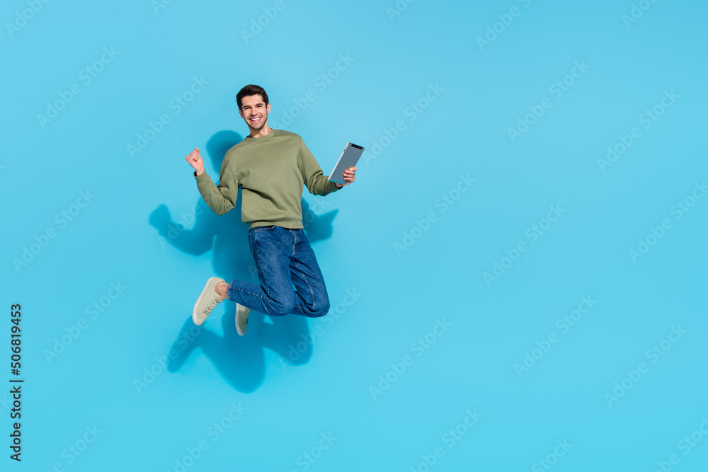 Full length photo of cool millennial brunet guy jump hold tablet wear shirt jeans shoes isolated on blue background Full length photo of cool millennial brunet guy jump hold tablet wear shirt jeans shoes isolated on blue background