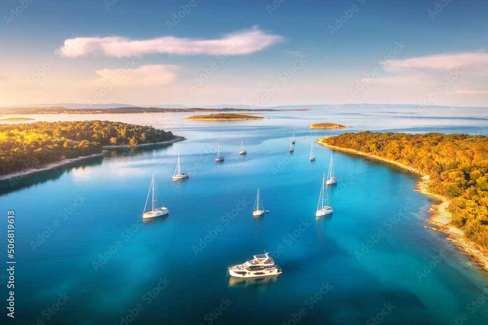 Aerial view of beautiful yachts and boats on the sea at sunset in summer. Adriatic sea, Kamenjak, Croatia. Top view of luxury yachts, sailboats, lagoon, clear blue water, and green forest. Travel - obrazy, fototapety, plakaty 