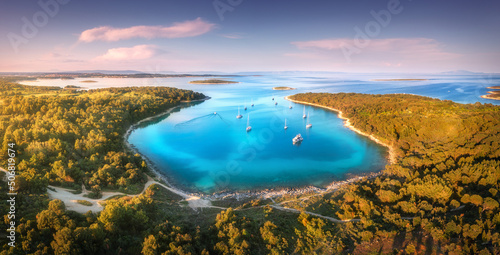 Fototapeta Naklejka Na Ścianę i Meble -  Aerial view of beautiful yachts and boats on the sea at sunset in summer. Adriatic sea, Kamenjak, Croatia. Top view of luxury yachts, sailboats, lagoon, clear blue water, and green forest. Travel