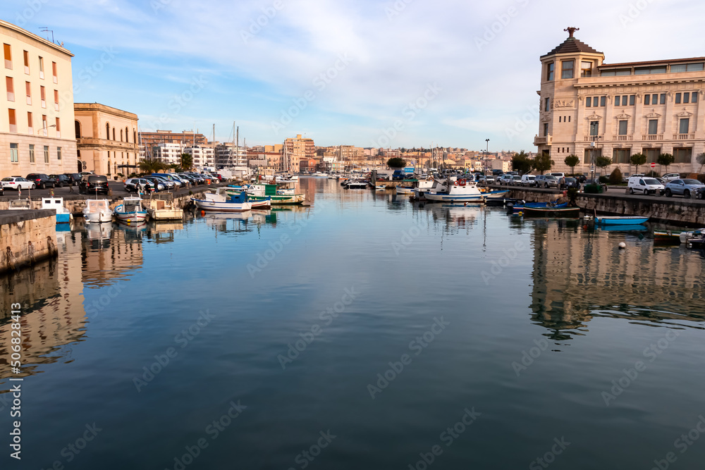 View of fishing boats moored in historic inner harbor in the city of Syracuse (Siracusa), Sicily, Italy, Europe, EU. Port connecting Syracuse with Ortygia Island. Reflection in water on a sunny day