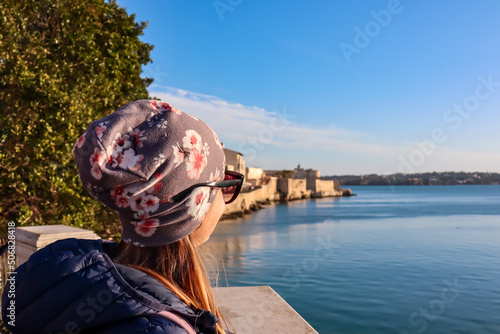 Tourist woman with panoramic view on waterfront of the city of Syracuse, Sicily, Italy, Europe EU. Soft light shining on residential houses at the Mediterranean seaside. Walking along the coastline